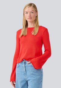 Red long-sleeve textured sweater with a square neckline and flared cuffs, worn with light blue high-waisted jeans.
