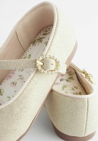 Bailarinas en dorado claro con un acabado texturizado de purpurina, que presentan una hebilla decorativa con rhinestones y un patrón floral en el interior.