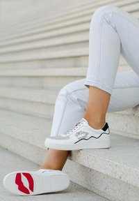 Baskets blanches avec des accents noirs et un ornement en forme de lèvres argenté, présentant une semelle texturée et un dessus lisse, posées sur des marches.