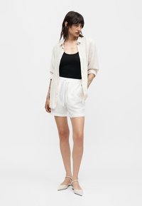 Γυναίκα που φοράει μαύρο αμάνικο τοπ, λευκό άνετο πουκάμισο με κουμπιά, λευκά σορτς με κορδόνι και λευκές μυτερές μπαλαρίνες, στέκεται με τα χέρια στις τσέπες.