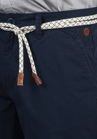 Navy blue shorts with a textured fabric, featuring a woven cream belt with a cross pattern and brown leather accents on the ends.