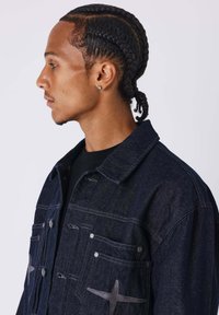 Giacca in denim scuro con collo a risvolto, dotata di due tasche frontali, accessori in argento e un distintivo ricamo sul petto.