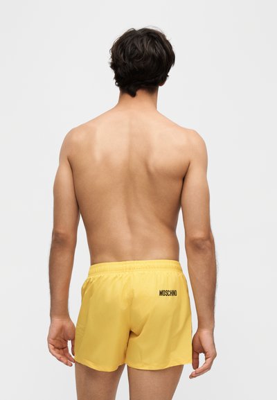 Homme aux cheveux courts et foncés, debout de dos, torse nu, portant un short jaune avec le logo Moschino sur le côté droit.