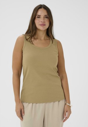 Kvinde med langt brunt hår iført en ærmeløs beige top og lyse bukser, stående mod en ensfarvet hvid baggrund.