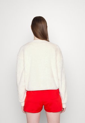 Femme aux longs cheveux bruns porte un pull blanc duveteux et un short rouge vif, debout face à un fond blanc uni.