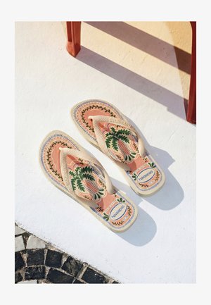 Paar beige Flip-Flops mit grünen Blattmustern und Markenlogo auf einer weißen Oberfläche neben einem roten Stuhlbein, das einen Schatten wirft.