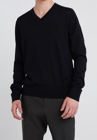Maglione nero a V in morbido tessuto a maglia, con maniche lunghe e polsini e orlo a coste per un aspetto aderente. Indossato sopra pantaloni scuri.