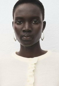 Cardigan bianco a coste con bordatura arricciata e chiusura con bottoni. La persona ha i capelli corti e indossa orecchini a cerchio d'argento su uno sfondo chiaro.