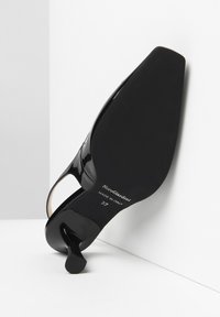 Scarpa con tacco alto in vernice nera, punta appuntita, schiena aperta e tacco sottile. Presenta una finitura liscia e un logo impresso sulla suola.