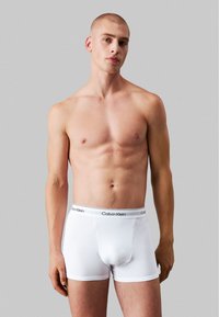 Boxer-briefs en coton blanc avec une ceinture noire à logo, design ajusté et longueur mi-cuisse sur un modèle sur fond gris.