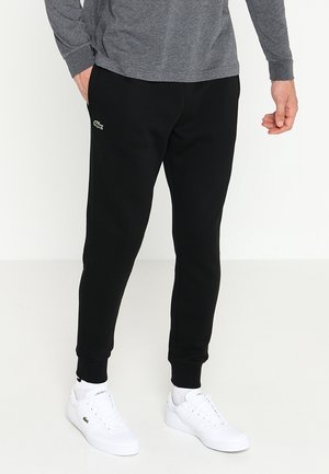 Mand iført sorte joggingbukser med Lacoste-logo på låret, hvide Lacoste-sneakers og grå langærmet trøje, stående på hvidt gulv.
