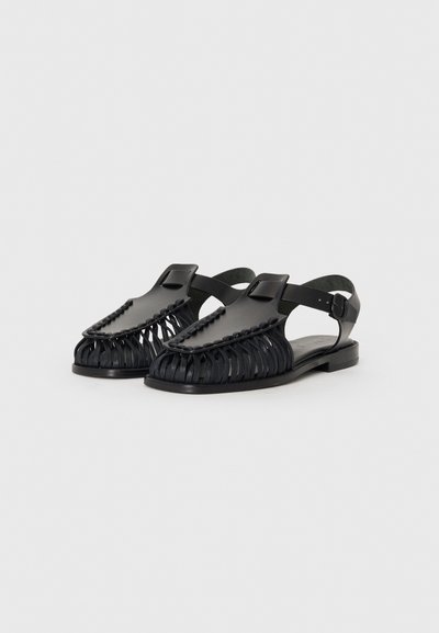 Scarpe slingback nere con tomaia in pelle intrecciata, chiusura con cinturino e fibbia, suola piatta e frange decorative sulla punta.
