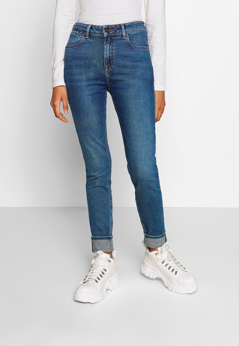 Jeans ajustados de color azul oscuro con cintura alta, con bolsillos traseros y un ligero desvanecimiento. Combinados con zapatillas gruesas blancas.