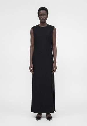 Femme portant une robe maxi noire sans manches avec des fentes sur les côtés et des chaussures plates pointues noires, debout devant un fond blanc uni.