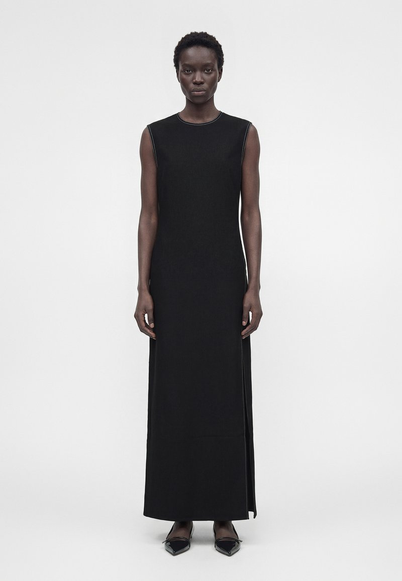 Γυναίκα που φοράει μαύρο μακρύ φόρεμα χωρίς μανίκια με ανοίγματα στο πλάι και μαύρα μυτερά μπαλαρίνια, στέκεται μπροστά σε απλό λευκό φόντο.