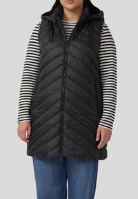 Schwarze Steppweste mit Kapuze, mit Chevron-Steppung, Reißverschluss und Seitentaschen, getragen über einem schwarz-weißen gestreiften Langarmshirt.