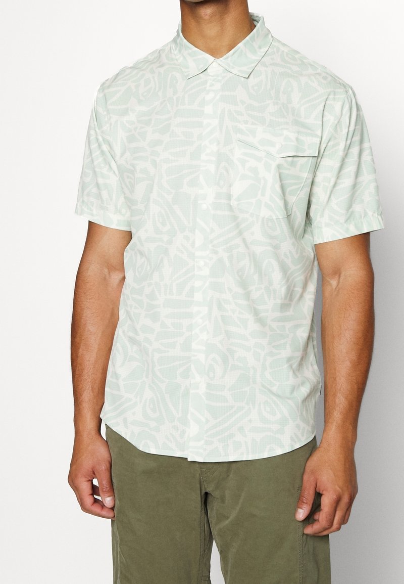 Homme portant une chemise à manches courtes avec un motif abstrait vert clair et blanc et un pantalon vert olive, se tenant devant un fond blanc.