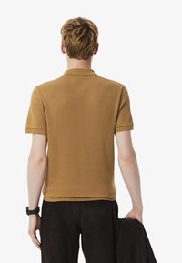 Camisa polo marrón con un corte relajado, de mangas cortas y un cuello sutil. El material parece suave. Combinada con pantalones oscuros, sosteniendo una chaqueta en la mano.