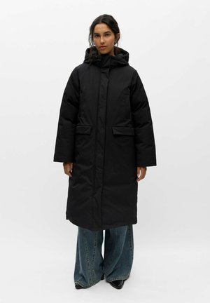Femme debout portant un long manteau d'hiver noir à capuche par-dessus un jean bleu à jambes larges et des chaussures noires sur un fond blanc uni.