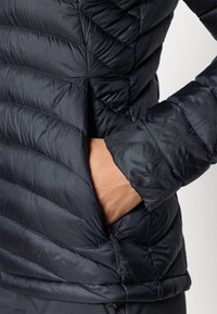 Czarna kurtka ocieplana z pikowaną strukturą i skośnym przeszyciem, z zamykaną na zamek kieszenią oraz miękkim, połyskującym wykończeniem.