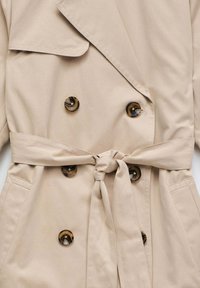 Manteau beige avec un design croisé, grandes boutons marron, ceinture à nouer et poches latérales. Tissu lisse avec une texture douce.
