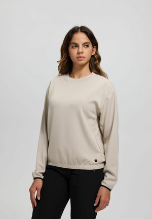 Giovane donna con capelli mossi che indossa una maglia beige a maniche lunghe e pantaloni neri, in piedi contro uno sfondo chiaro e uniforme.