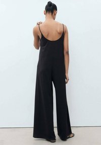 Mono negro con espalda al aire, tirantes finos y piernas anchas. Fabricado en una tela suave, presenta un ajuste relajado y un diseño minimalista.