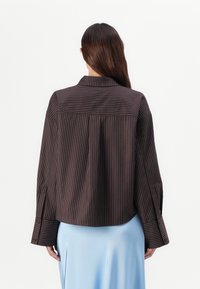 Chemise marron à rayures avec un col et des poignets boutonnés, présentant une coupe décontractée et un design cropped, associée à une jupe bleu clair.
