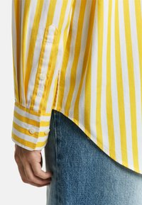 Camicia a strisce gialle e bianche con polsini abbottonati, caratterizzata da un orlo curvo e realizzata in morbido cotone, abbinata a jeans di denim blu.