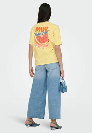 Kvinde med langt mørkt hår iført gul T-shirt med pink grapefrugtgrafik, brede blå jeans, turkis slingback-højhælede sko og bærende en lille blå håndtaske.