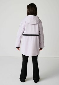 Svetlofialová nepremokavá bunda s kapucňou, čiernymi akcentmi a sťahovacím pásom. Má dlhé rukávy a zakrivený lem, v kombinácii s čiernymi nohavicami.