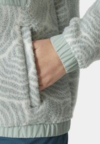 Jasnoszary polarowy sweatshirt o teksturowanej powierzchni; ma kieszeń na zamek i elastyczne mankiety; dolny brzeg wykonany jest z gładkiego, miętowozielonego materiału.