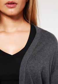 Šedý kardigan vyrobený z pleteného materiálu, s otevřeným předním dílem a měkkou texturou přes černé v-neck tričko. Vzhled zblízka.