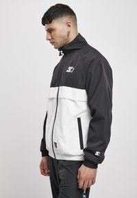 Giacca zip-up bianca e nera con colletto alto, polsini elastici, tasche laterali e un logo a stella sul petto. Tessuto dalla consistenza liscia.