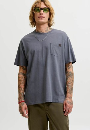 Jeune homme aux cheveux blonds et tatouages porte un t-shirt gris avec une poche, un pantalon olive, des lunettes de soleil vertes, un collier en argent et des bracelets rouges.