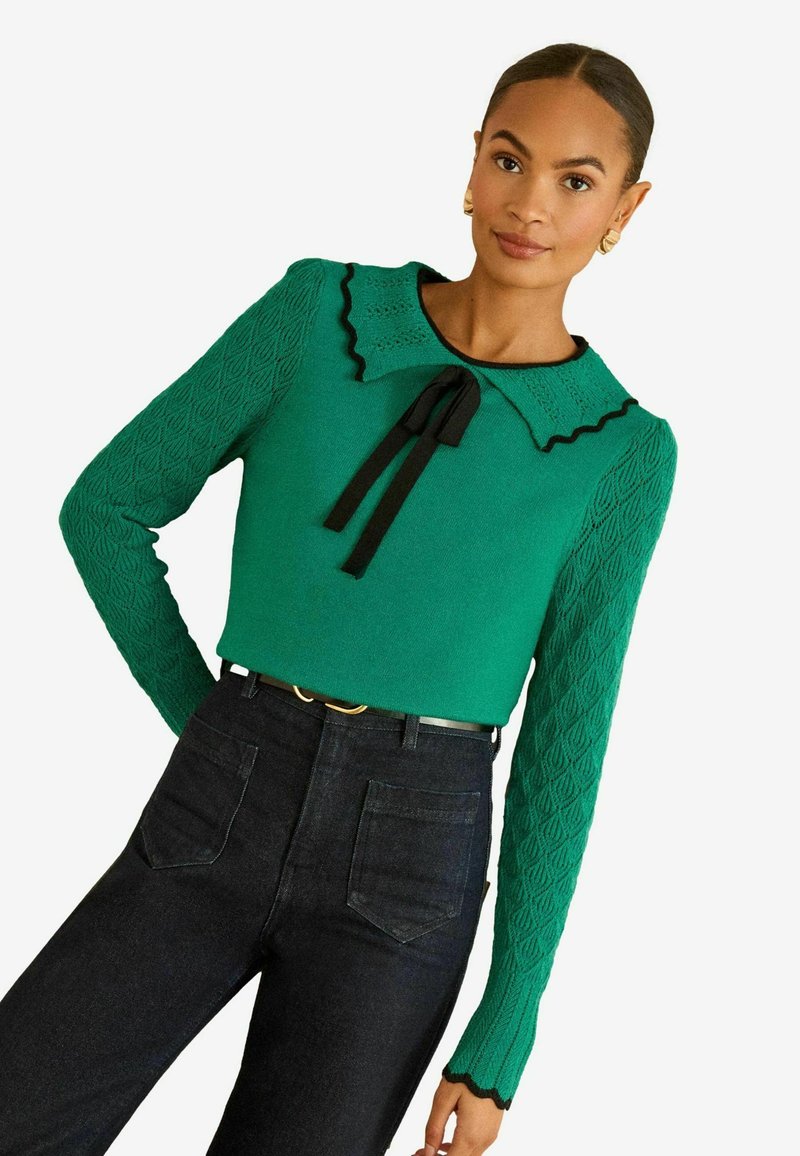 Grøn sweater med teksturerede, mønstrede ærmer, sort bølget kant og en sløjfe ved halsudskæringen, kombineret med mørke denim højtaljede jeans.