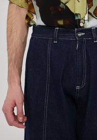 Tmavé džínsové nohavice s kontrastným bielym prešívaním, s rovnými nohavicami. Obsahujú predné zapínanie na gombík a bočné vrecká.