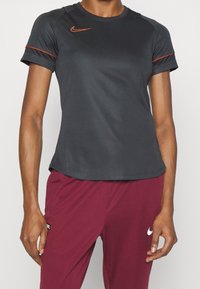 Person som bär en svart t-shirt med kort ärm från Nike med en orange logotyp och maroon Nike-byxor mot en enfärgad vit bakgrund.