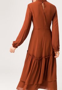 Nainen, joka on pukeutunut pitkähihamaiseen, ruosteenväriseen mekkoon, jossa on röyhelöhihet ja kerroksellinen hame, sidottuna rusetilla takana kaula-aukossa.