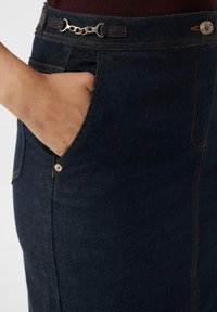 Hand in die vordere Tasche eines dunkelblauen Jeansrocks mit orangefarbenen Nähten und kleinen Metallkettendetails am Taillenbund.