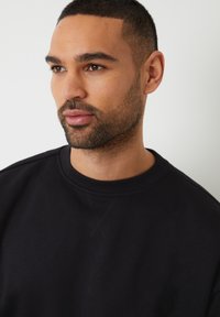 Zwarte sweatshirt met ronde hals, met een gladde textuur en zichtbare stiksel-details op de voorkant en schouders.