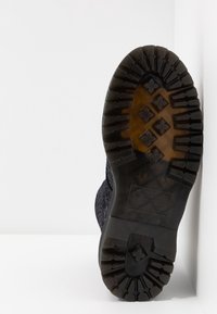 Semelle d'une botte noire avec des crampons profonds et robustes reposant verticalement contre un mur d'angle blanc sur une surface blanche.