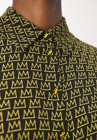 Camicia a maniche corte con sfondo nero e motivo a ripetizione di corone gialle, completata da bottoni gialli a contrasto nel colletto.