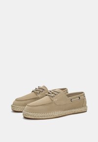 Beige vászoncipő, lekerekített orral, fűzős elülső résszel és espadrille-stílusú kender talppal. Fém fűzőlyukakkal és varrási részletekkel rendelkezik.