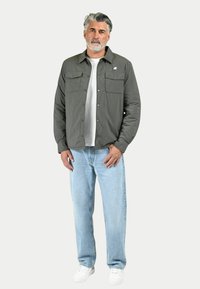Giacca verde oliva da uomo con due tasche sul petto, indossata sopra una tshirt bianca, abbinata a jeans blu chiaro e sneakers bianche.