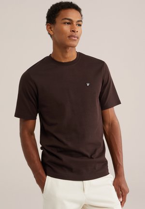 Uomo in piedi con le mani in tasca, indossa una maglietta a maniche corte marrone scuro e pantaloni beige chiaro su uno sfondo semplice.
