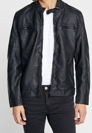 Homme portant une veste en cuir noire avec des poches zippées sur la poitrine et les côtés, par-dessus un t-shirt blanc et un pantalon noir.