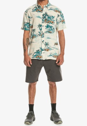 Mann, der ein kurzärmeliges Hemd mit tropischem Muster, dunkle Shorts, schwarze Socken und graue Sportschuhe trägt, steht vor einem weißen Hintergrund.