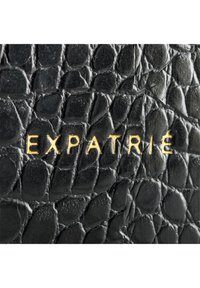 Schwarzes strukturiertes Leder mit einem Reptilleder-Muster, mit dem Markennamen "EXPATRIÉ" in Gold geprägt.