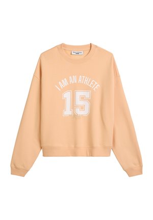 Bluzka z długim rękawem w kolorze brzoskwiniowym, ze ściągaczami u rękawów i okrągłym dekoltem. Na przodzie znajduje się biała grafika z napisem: "JESTEM SPORTOWCEM" oraz wyraźnie widoczną cyfrą "15".